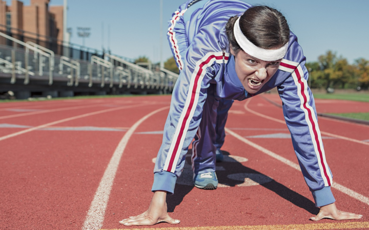 Mujer atleta corriendo en una pista de ceniza, en la salida de un sprint, con ropa deportiva y cinta en la cabeza.
