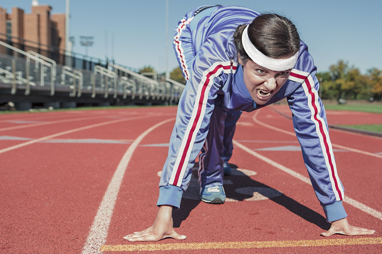 Mujer atleta corriendo en una pista de ceniza, en la salida de un sprint, con ropa deportiva y cinta en la cabeza.