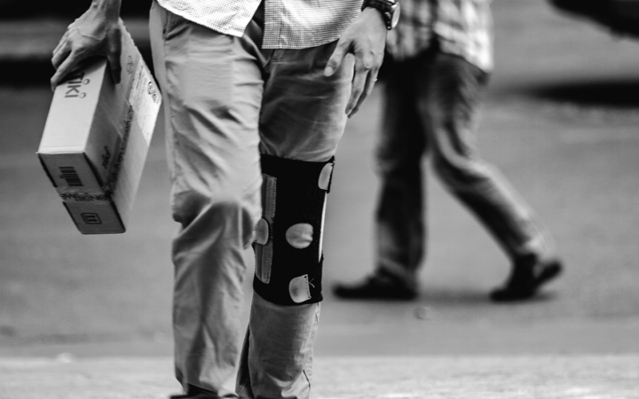 Persona en blanco y negro, con piernas apoyadas en una órtesis, caminando al aire libre mientras carga un paquete con ambas manos.