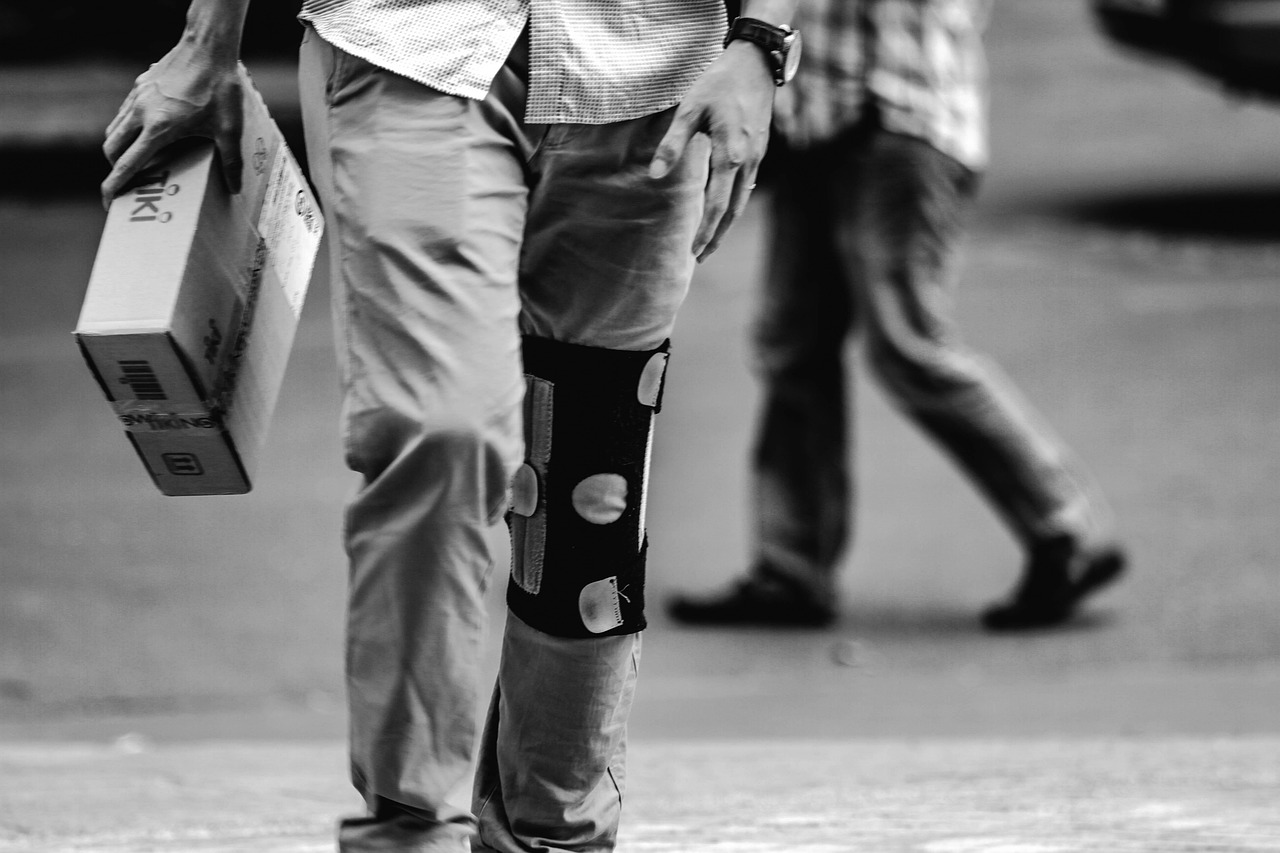 Persona en blanco y negro, con piernas apoyadas en una órtesis, caminando al aire libre mientras carga un paquete con ambas manos.