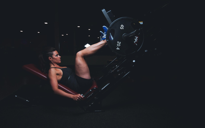 Mujer adulta y atlética entrenando en un gimnasio oscuro, con equipo de ejercicio y pesas, con piernas musculosas y gesto de esfuerzo.